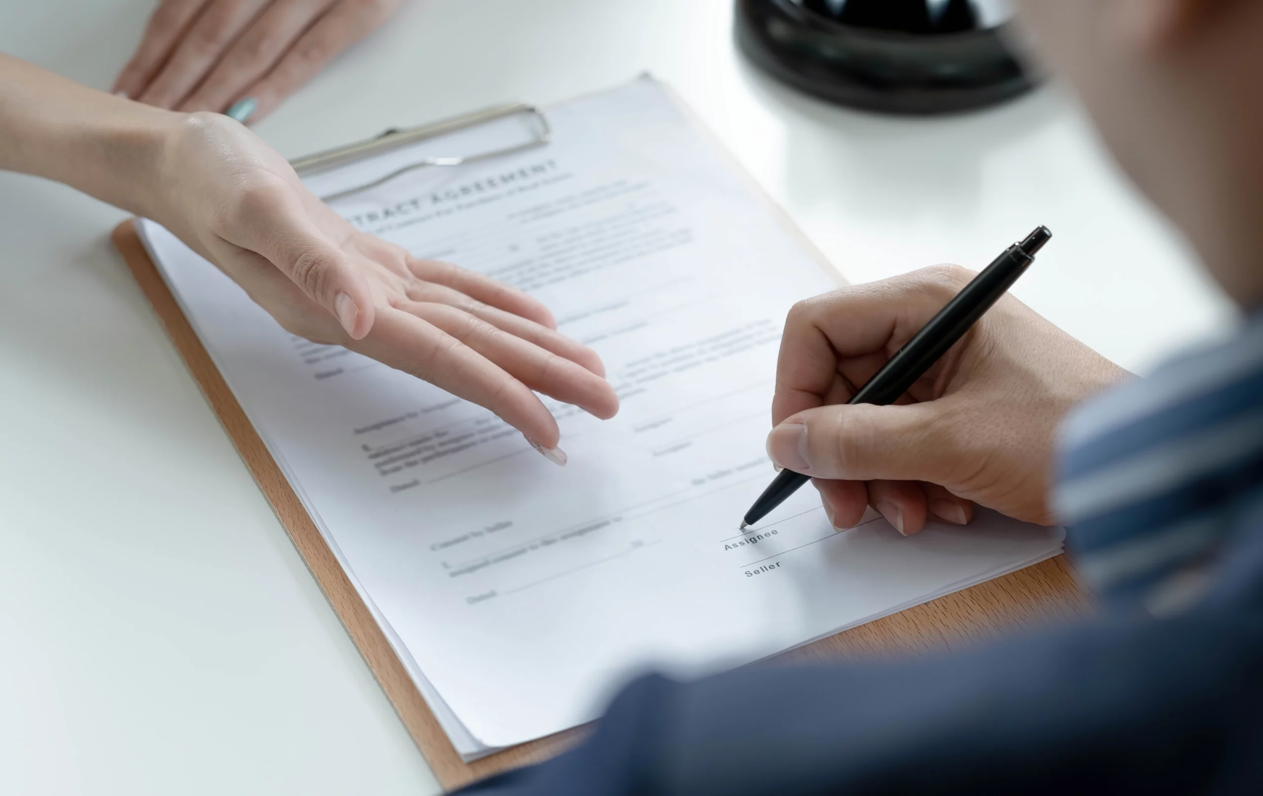 Person signing a contract on a clipboard with a pen while another hand gestures nearby, symbolizing legal agreement.